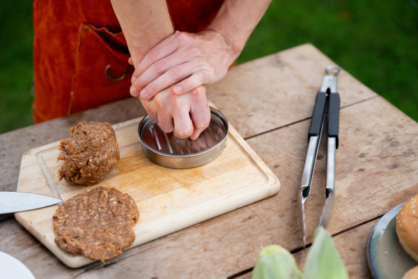 Man in apron preparing meat for hamburgers, standing outdoors. Using burger press to shape groud meat.