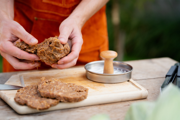 Man in apron preparing meat for hamburgers, standing outdoors. Using burger press to shape groud meat.