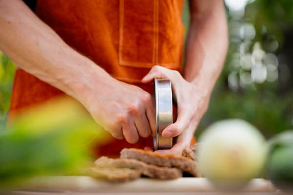 Man in apron preparing meat for hamburgers, standing outdoors. Using burger press to shape groud meat.