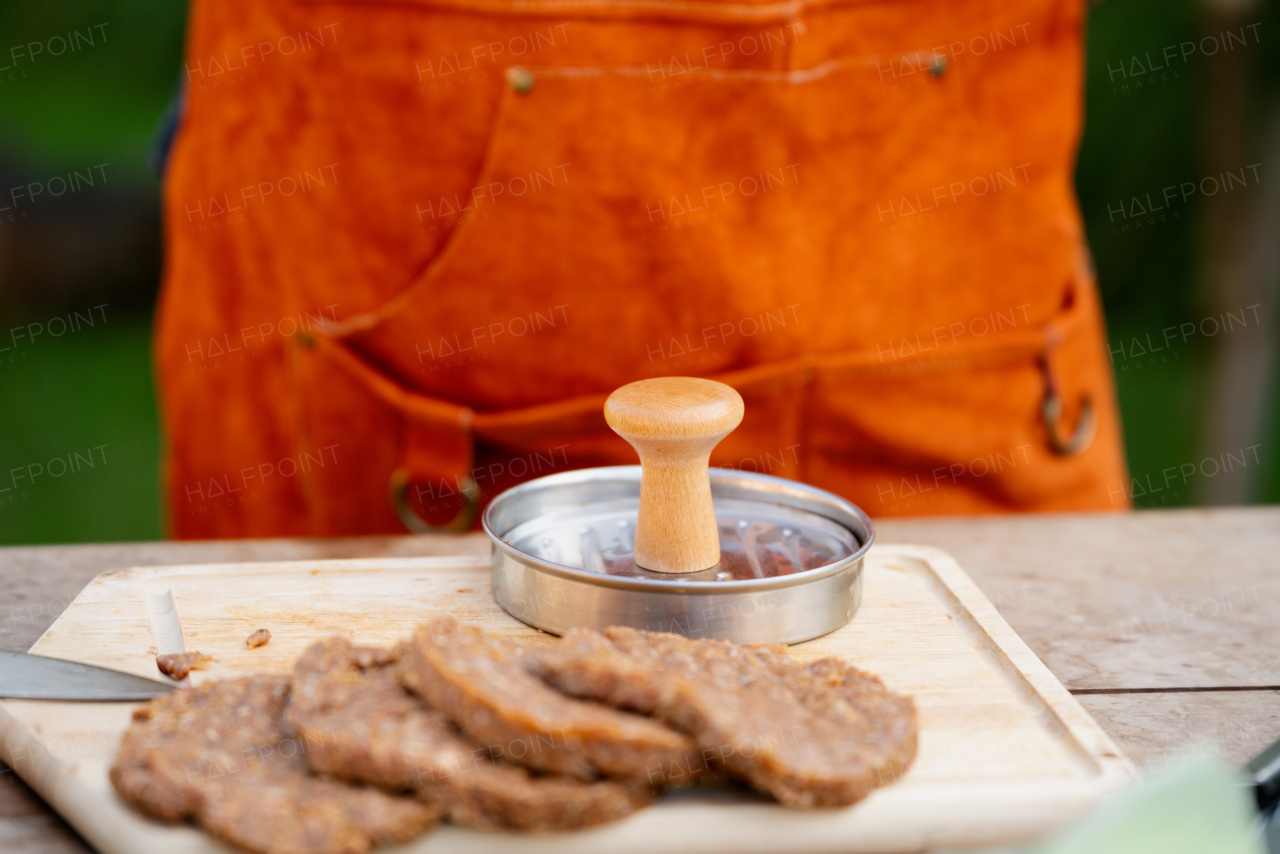 Man in apron preparing meat for hamburgers, standing outdoors. Using burger press to shape groud meat.