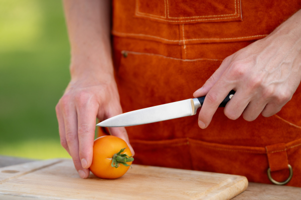 Close up of man holding sharp knife, slicing juicy red tomato. Preparing vegetables for outdoor barbecue.