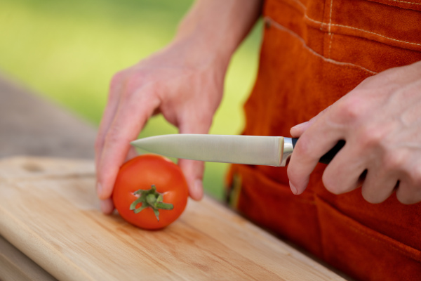 Close up of man holding sharp knife, slicing juicy red tomato. Preparing vegetables for outdoor barbecue.