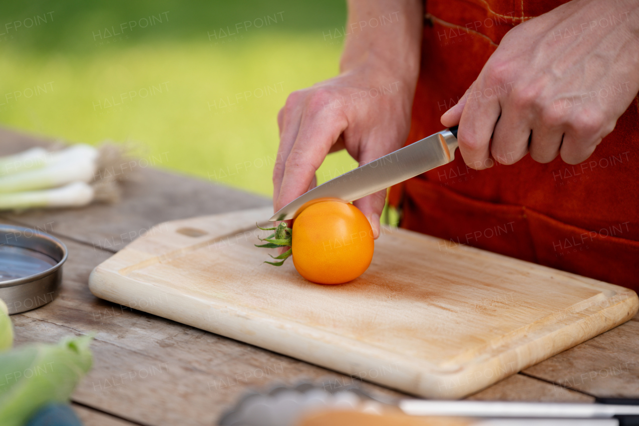 Close up of man holding sharp knife, slicing juicy red tomato. Preparing vegetables for outdoor barbecue.