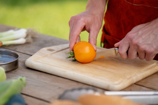 Close up of man holding sharp knife, slicing juicy red tomato. Preparing vegetables for outdoor barbecue.