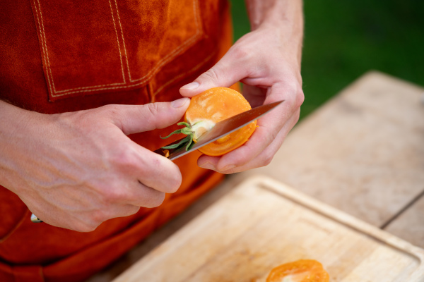 Close up of man holding sharp knife, slicing juicy red tomato. Preparing vegetables for outdoor barbecue.