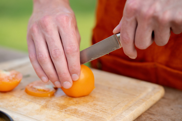 Close up of man holding sharp knife, slicing juicy red tomato. Preparing vegetables for outdoor barbecue.