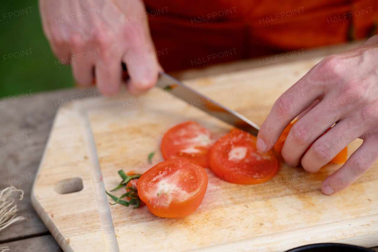 Close up of man holding sharp knife, slicing juicy red tomato. Preparing vegetables for outdoor barbecue.