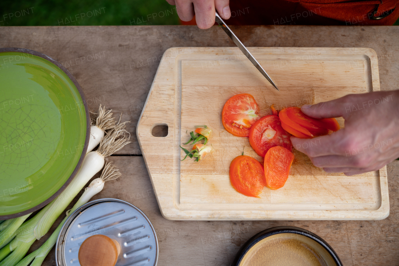 Close up of man holding sharp knife, slicing juicy red tomato. Preparing vegetables for outdoor barbecue.