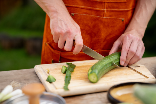 Close up of man holding sharp knife, peeling salad cucumber. Preparing vegetables for an outdoor barbecue.