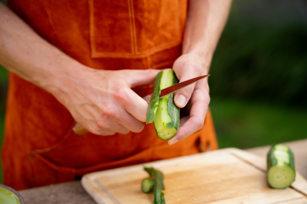 Close up of man holding sharp knife, peeling salad cucumber. Preparing vegetables for an outdoor barbecue.