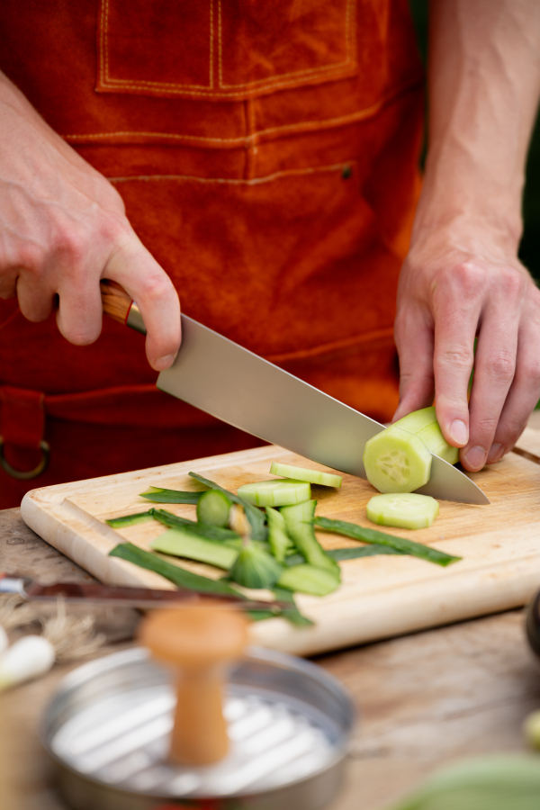 Close up of man holding sharp knife, chopping salad cucumber. Preparing vegetables for an outdoor barbecue.