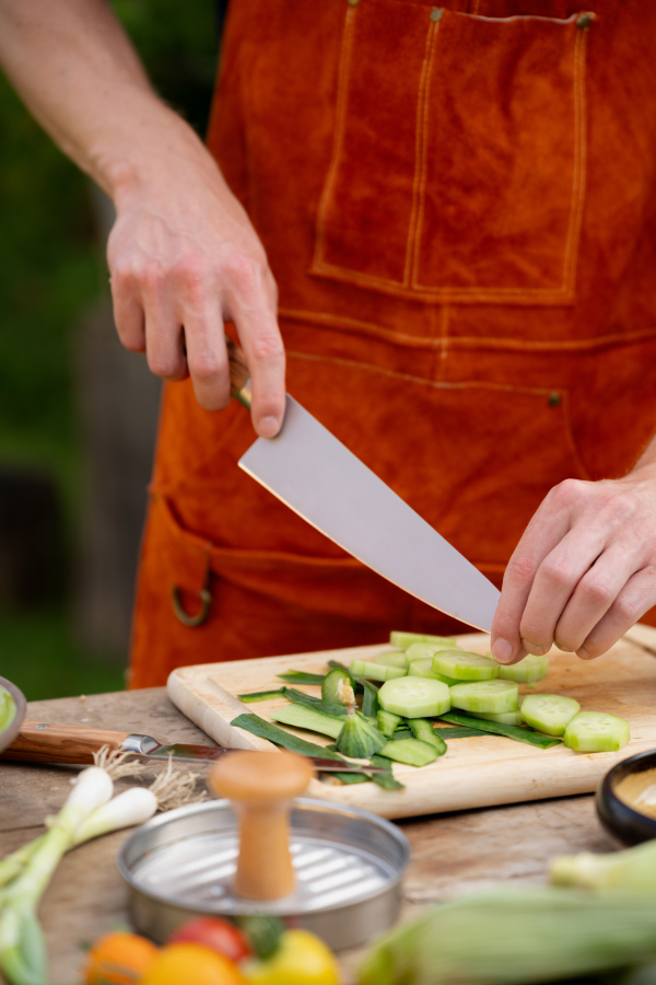 Close up of man holding sharp knife, chopping salad cucumber. Preparing vegetables for an outdoor barbecue.