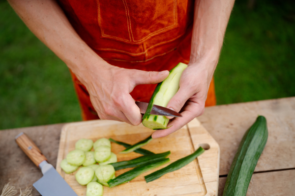 Close up of man holding sharp knife, peeling salad cucumber. Preparing vegetables for an outdoor barbecue.