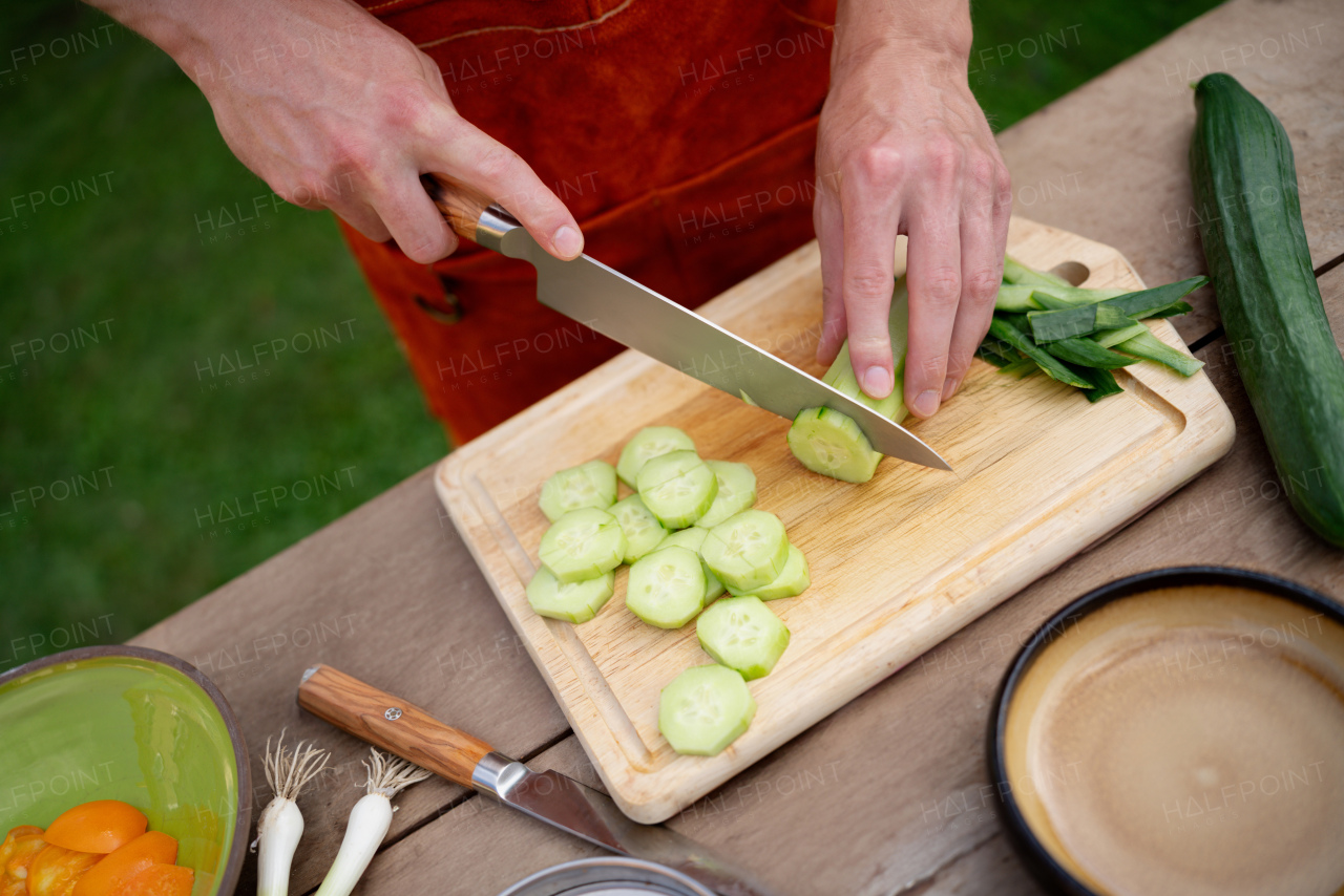 Close up of man holding sharp knife, chopping salad cucumber. Preparing vegetables for an outdoor barbecue.