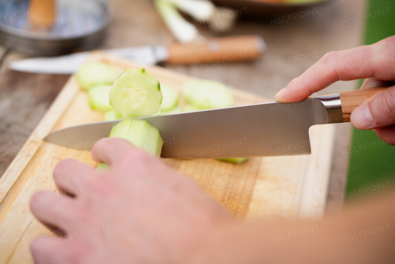 Close up of man holding sharp knife, chopping salad cucumber. Preparing vegetables for an outdoor barbecue.