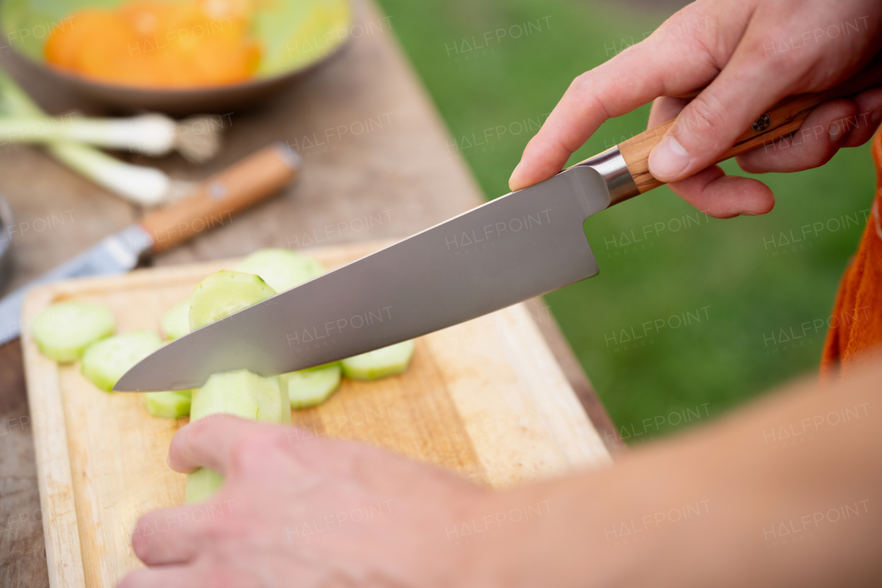 Close up of man holding sharp knife, chopping salad cucumber. Preparing vegetables for an outdoor barbecue.