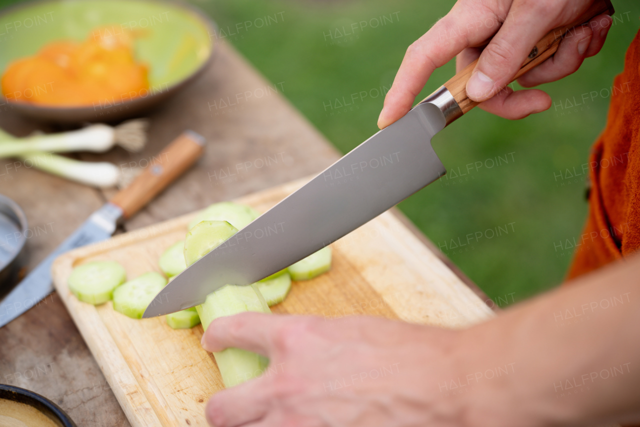 Close up of man holding sharp knife, chopping salad cucumber. Preparing vegetables for an outdoor barbecue.