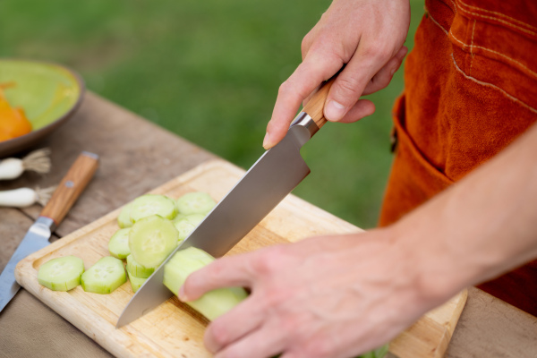 Close up of man holding sharp knife, chopping salad cucumber. Preparing vegetables for an outdoor barbecue.