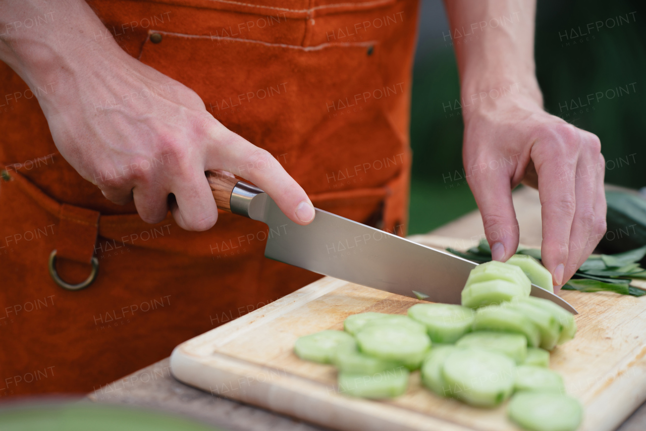 Close up of man holding sharp knife, chopping salad cucumber. Preparing vegetables for an outdoor barbecue.