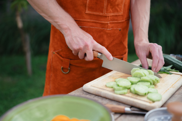 Close up of man holding sharp knife, chopping salad cucumber. Preparing vegetables for an outdoor barbecue.