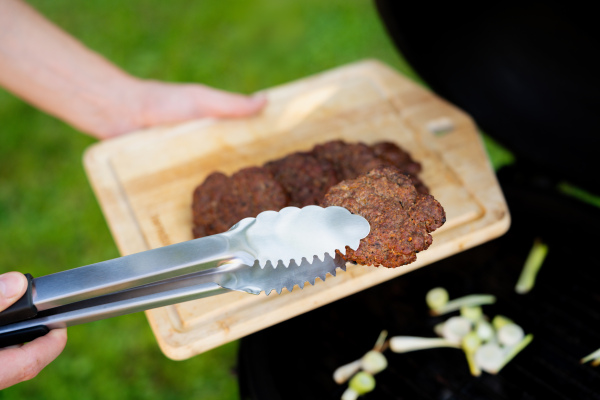 Close upf of man serving grilled burger patties from cutting board with sharp, using grill tongs. Preparing food for an outdoor barbecue.