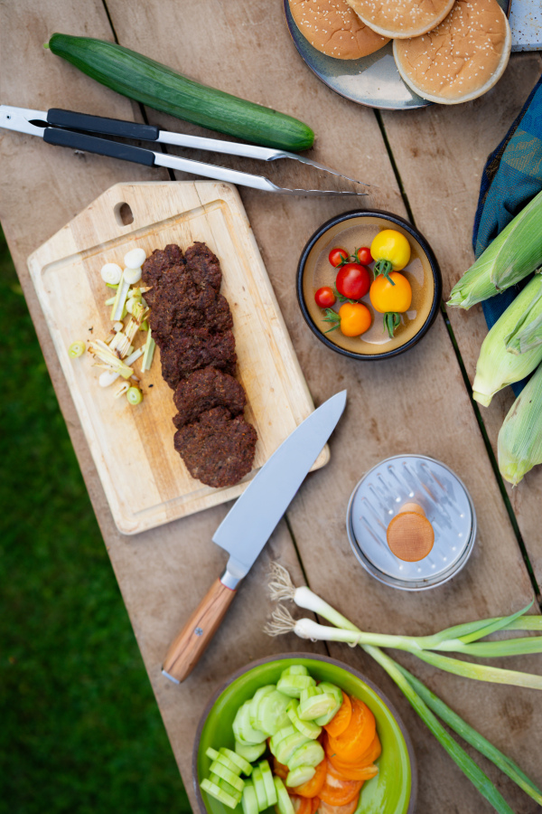 Top view of cutting board with sharp knife, chopped vegetables and grilled burger patties. Preparing food for an outdoor barbecue.