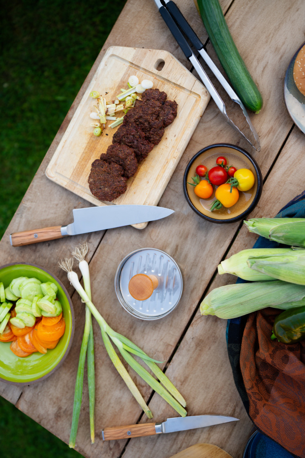 Top view of cutting board with sharp knife, chopped vegetables and grilled burger patties. Preparing food for an outdoor barbecue.