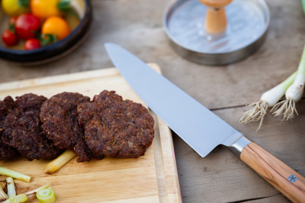 Cutting board with sharp knife, vegetables and grilled burger patties. Preparing food for an outdoor barbecue.