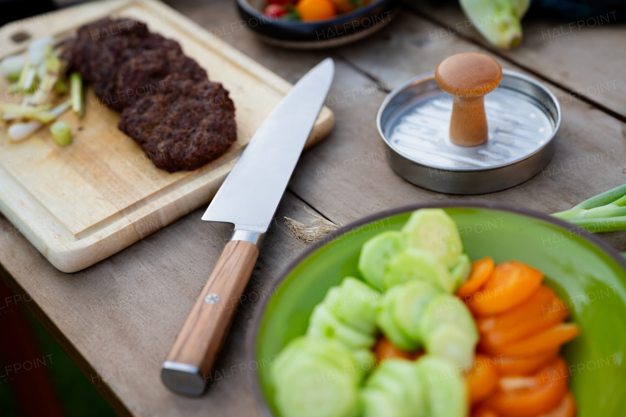 Preparing food for an outdoor barbecue. Cutting board with sharp knife, chopped vegetables, hamburger patty press and grilled burger patties.