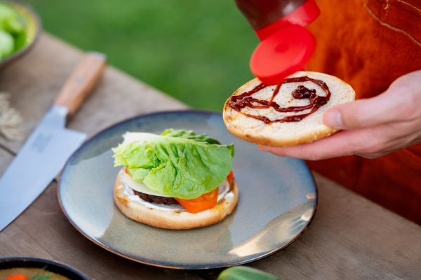 Close up of man assembling burgers with toppings, putting barbecue sauce on toasted bun.