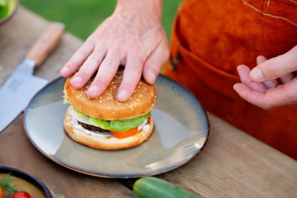 Close up of man assembling burgers with toppings, putting toasted bun on top of hamburger.