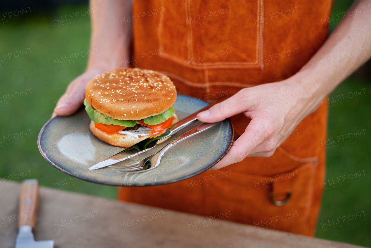 Close up of man holding plate with homemade grilled hamburger and cutlery.