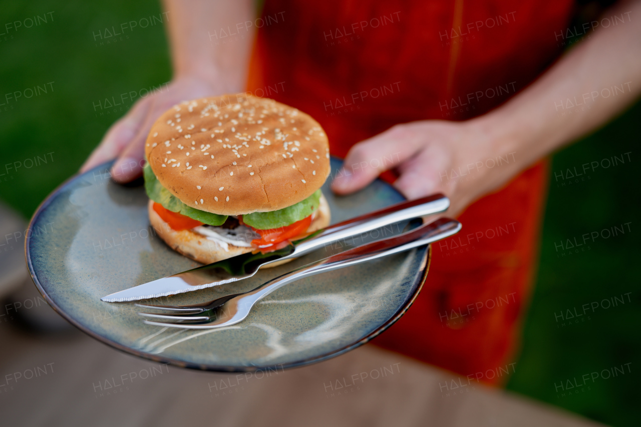 Close up of man holding plate with homemade grilled hamburger and cutlery.