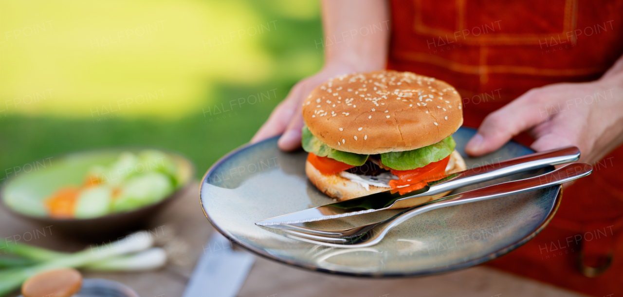 Close up of man holding plate with homemade grilled hamburger and cutlery. Banner with copy space.