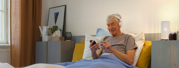 Senior man lying in a bed and enjoying music trough headphones.