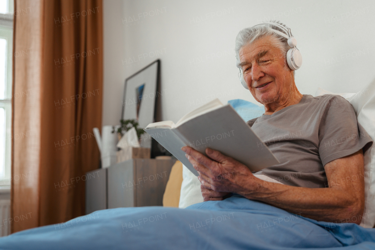 Senior man relaxing in his bed, learning book and listening music through headphones