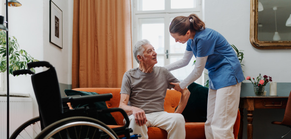 Young nurse helping senior man to stand up from a sofa.