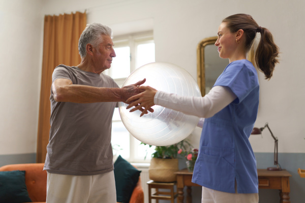 Young nurse doing exercise with senior man in his home. Young physiotherapist helping senior man to do exercise with fit ball, gymanstic ball.