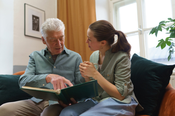 Grandfather with his granddaughter looking at family photo album. Nostalgic senior man showing photos of himself from his youth, remebering old days through pictures.