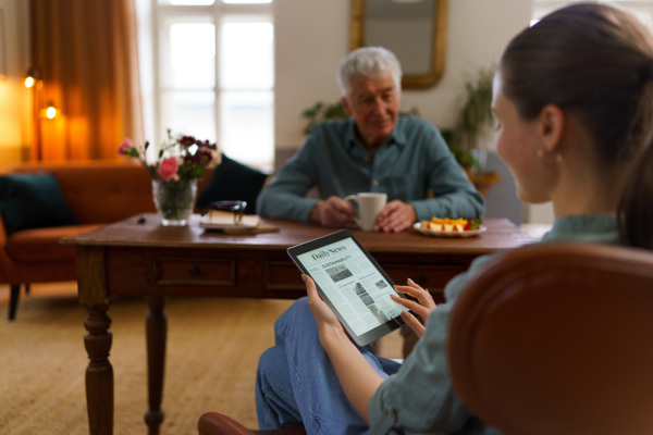 Caregiver reading online newspaper in digital tablet during taking care of senior man in his home.