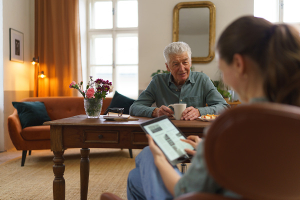 Caregiver reading online newspaper in digital tablet during taking care of senior man in his home.