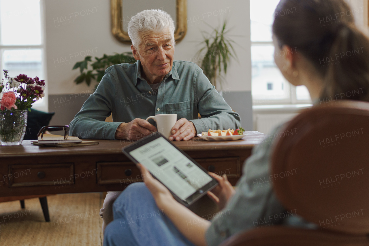 Caregiver reading online newspaper in digital tablet during taking care of senior man in his home.