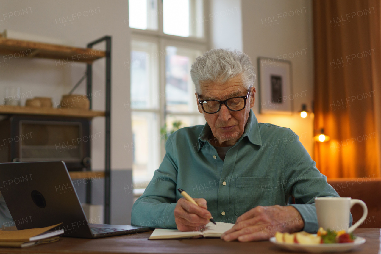 Senior man writing notes in his diary, with opened computer.