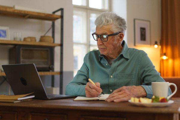 Senior man writing notes in his diary, with opened computer. Elderly man eating snack in front of a computer.