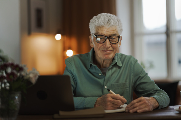 Senior man writing notes in his diary, with opened computer.