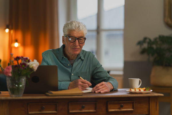 Senior man writing notes in his diary, with opened computer.