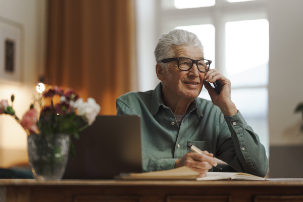 Senior man calling and writing some notes in his apartment.