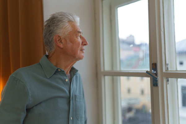 Portrait of handsom elderly man with gray hair. Senior man looking out of a window, concept of solitude in retirement.