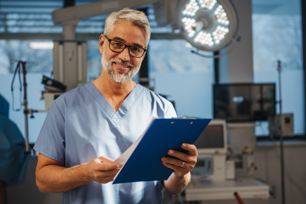 Portrait of confident ER doctor standing in hospital, emergency room. Handsome doctor in scrubs holding clipboard with test results, standing in modern private clinic, looking at camera.