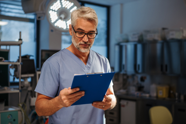 Portrait of confident ER doctor standing in hospital, emergency room. Handsome doctor in scrubs holding clipboard with test results, standing in modern private clinic, looking at camera.